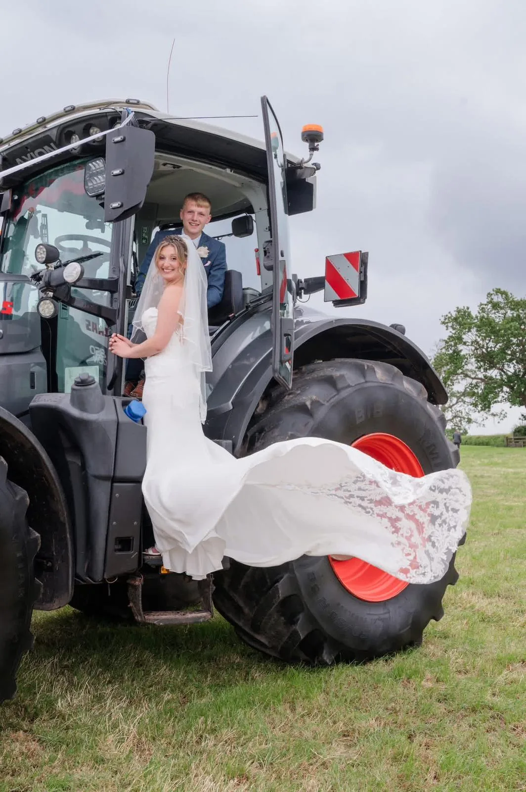 Bride and groom on a tractor on their wedding day, dress train flowing across the wheel
