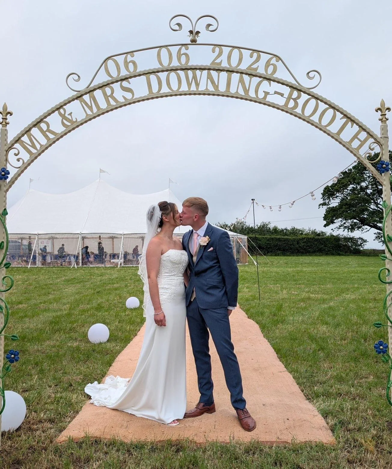 Bride and groom kissing beneath a countryside wedding arch in front of a marquee