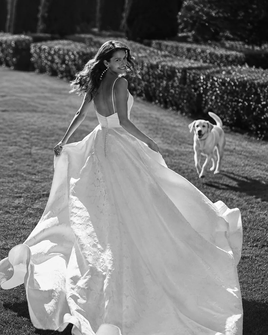 Bride in a flowing A-line wedding dress walking through a formal country garden with a Labrador running behind her, photographed in black and white — BOE Bridal, Eccleshall